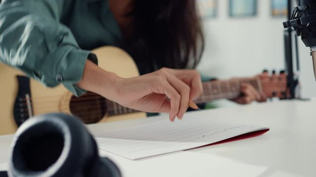 Happy asia woman songwriter play acoustic guitar listen song from smartphone think and write notes lyrics song in paper sit in living room at home studio. Music production at home concept.