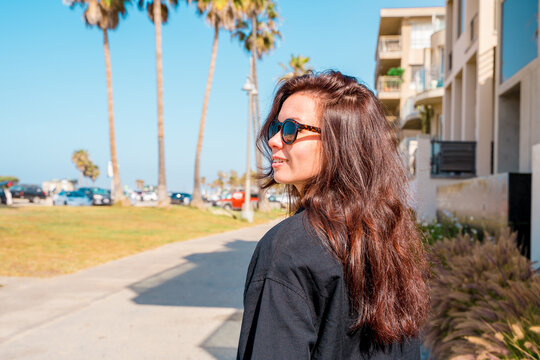 Cute young brunette girl in sunglasses walks along the Los Angeles waterfront with palm trees and streets on a summer day
