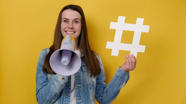Surprised young woman 20s screaming in megaphone showing white hashtag sign, wears denim jacket, isolated on yellow background. Marketing network, SMM business, customer attraction campaign concept