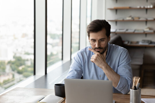 Thoughtful young male employee or worker sit at desk in office work online on laptop solve business problem. Pensive businessman use computer thinking pondering over project. Planning concept.