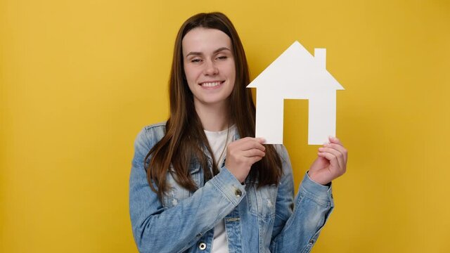 Charming positive young caucasian woman holding paper house,  international day of families, foster home care, isolated over yellow studio background with copy space. Home protection insurance concept