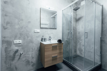 Contemporary bathroom with shower and sink with wooden cabinet under it and mirror with integrated light. All walls are painted with concrete trowel in gray. Whole space gives modern impression.