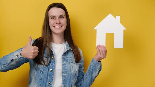 Charming young woman hold house model showing thumb up satisfied, cute female make like gesture recommending, satisfied with choice or decision, isolated on yellow wall. Housing and mortgage concept