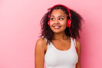 Young african american woman listening to music with headphones isolated on pink background looks aside smiling, cheerful and pleasant.