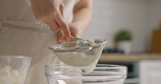 Woman in gray apron rubs cottage cheese with sieve at home kitchen. Close-up, slow motion