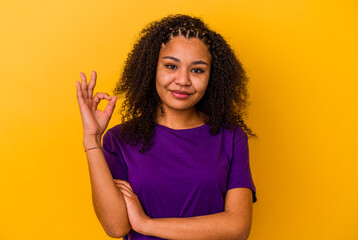 Young african american woman isolated on yellow background winks an eye and holds an okay gesture with hand.