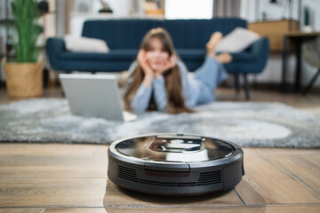 Black robot vacuum cleaner washing floor while caucasian woman with modern laptop relaxing on...