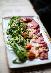 Delicious raw carpaccio served in restaurant on trendy white craft plate with green salad. Tomatoes and tasty dressing. High end dining. Healthy dish. Top view, selective focus