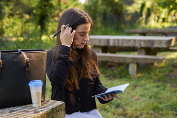 long-haired, fair-skinned pretty woman in black shirt thinking what to write in her notebook sitting at a stone table with coffee in nature with sunset light. smiling teenager looking for inspiration