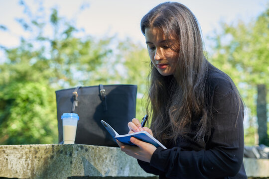long-haired pretty woman sitting on a stone table in nature writing in a notebook with coffee and her bag. teenager drawing in a blue notebook and black pen in the forest. girl making notes in agenda