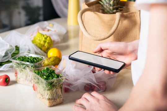 Online Home Food Delivery Of Fresh Vegetables And Fruits. Young Man Holding Phone And Checking Order List. Reusable Bag With Bio Vegetables On White Kitchen Table. Local Farmer Healthy Food.