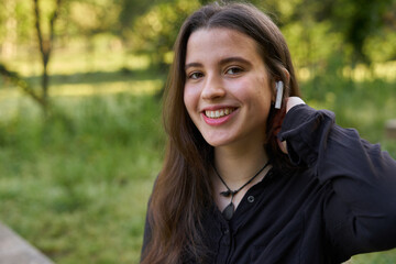 Naklejka premium long-haired, fair-skinned woman putting on white wireless headphones in nature. teenager using bluetooth audio devices with green blur background. girl listening to music in the park