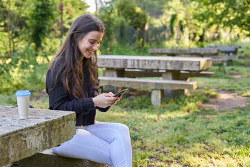 long-haired, fair-skinned woman sitting at a stone table with a coffee and her bag while talking on the phone, with green unfocused background. teenager in nature with his mobil taking a break.