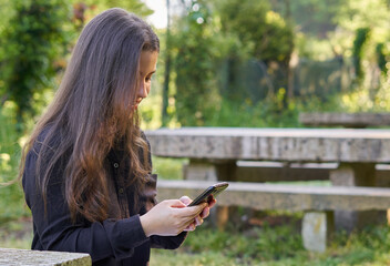 long-haired, fair-skinned woman sitting at a stone table with a coffee and her bag while talking on the phone, with green unfocused background. teenager in nature with his mobil taking a break.