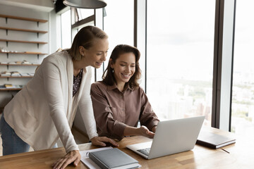 Smiling young female employees work together on computer at meeting in office. Happy women colleagues coworkers brainstorm on laptop discuss company financial business project. Teamwork concept.