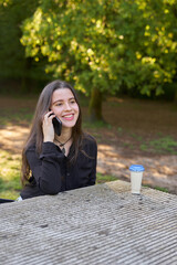 long-haired, fair-skinned woman sitting at a stone table with a coffee and her bag while talking on the phone, with green unfocused background. teenager in nature with his mobil taking a break.