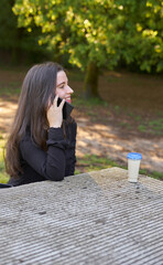 long-haired, fair-skinned woman sitting at a stone table with a coffee and her bag while talking on the phone, with green unfocused background. teenager in nature with his mobil taking a break.