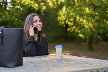 long-haired, fair-skinned woman sitting at a stone table with a coffee and her bag while talking on the phone, with green unfocused background. teenager in nature with his mobil taking a break.