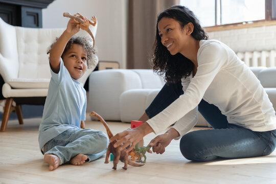 Happy cute kid boy and his mom relaxing on heat floor in cozy living room and playing with small dinosaurs figures together. Mother engaged in little sons game with toys. Family playtime concept