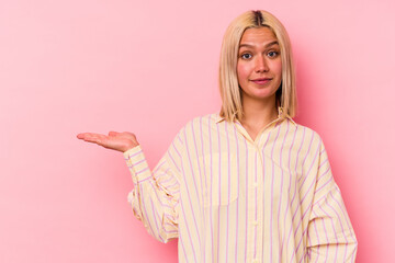 Young venezuelan woman isolated on pink background showing a copy space on a palm and holding another hand on waist.