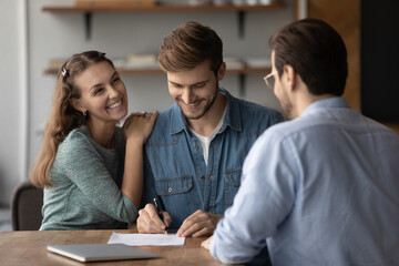 Overjoyed young couple buyers or client sign paper contract with male consultant at meeting. Happy man and woman male deal close agreement put signature on document with realtor in office.