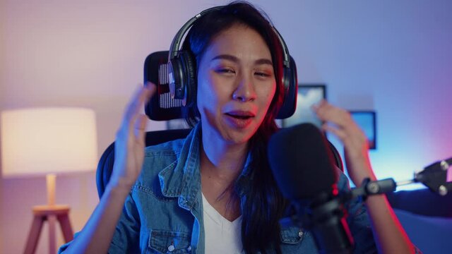 Happy asia girl blogger music influencer looking at camera broadcast record wear headphone online live talk in microphone with audience in living room home studio at night. Content creator concept.