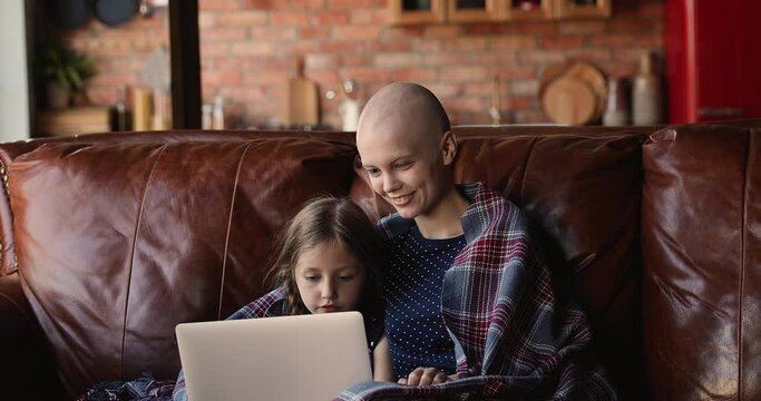 Happy young woman with oncology remission resting on comfortable sofa with smiling adorable kid daughter, watching funny movie or cartoons online on computer, using applications, playing games.
