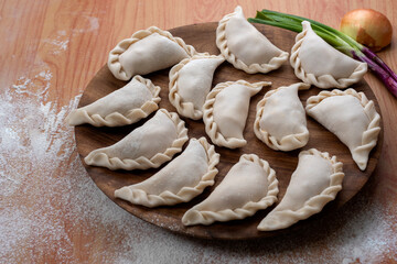 Argentine raw empanadas on round wooden plate on wooden table top with flour decorated with vegetables