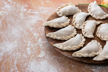 Argentine raw empanadas on round wooden plate on wooden table top with flour decorated with vegetables