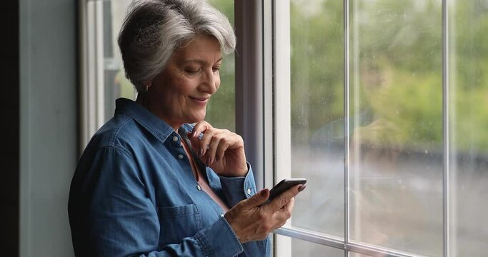 Smiling dreamy elderly senior woman standing near window, using smartphone applications, enjoying communicating distantly with grown children or friends, reading message with pleasant news indoors.