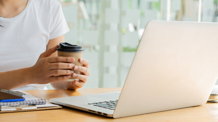 Young business woman holding a paper cup while working with laptop in a coffee shop.