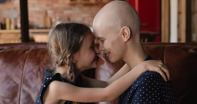 Sincere smiling little child girl touching noses with affectionate hairless after chemotherapy mother, showing love and devotion. Happy cute small kid daughter supporting mum with cancer illness.