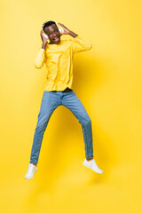 Fototapeta na wymiar Full length portrait of happy young African man wearing headphones listening to music and jumping in yellow isolated studio background