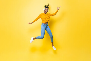 Fototapeta na wymiar Happy energetic young African American woman wearing headphones listening to music and jumping with hand up in yellow isolated studio background