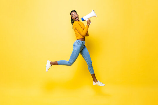 Jumping portrait of shocked young African American woman holding megaphone making announcement in isolated studio yellow background