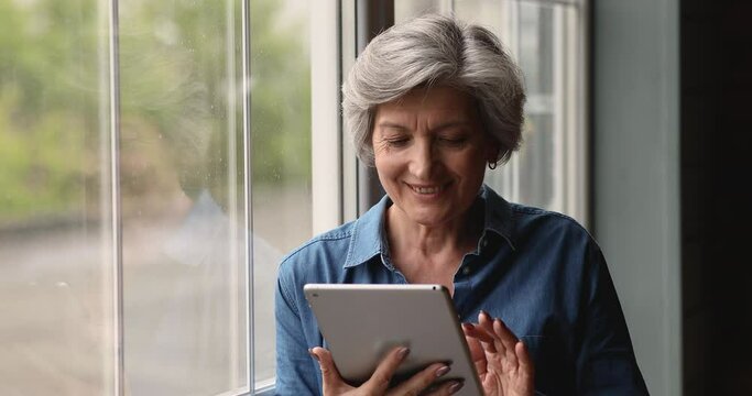 Smiling 60s older woman looking at digital tablet screen, watching family photo or video, choosing goods in internet store, shopping online or using software applications, standing near window.
