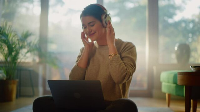 Young Woman Using Laptop at Home, Does Remote Work, Listens Music through Headphones. Beautiful Smiling Girl Sitting on the Floor Enjoys Music, Dances a Bit, Brainstorms Creative Project