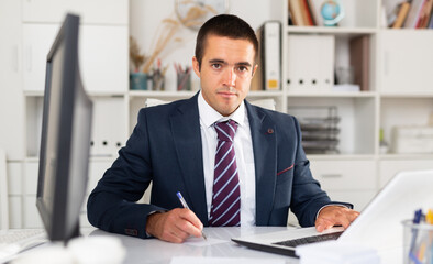 Obraz premium Portrait of successful busy entrepreneur sitting at office desk with papers and laptop