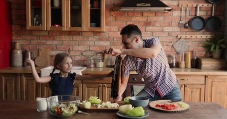 Joyful active little 7s kid girl playing battle with young father, fighting using kitchenware utensils. Happy different generations family having fun, entertaining distracted form cooking in kitchen.
