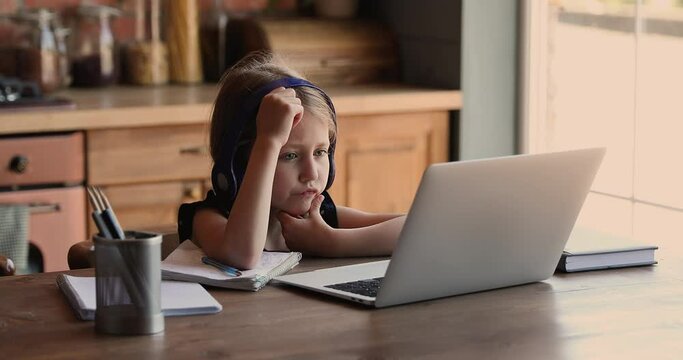 Concentrated little school aged cute girl in headphones web surfing information online, preparing homework, listening education lecture, writing notes, involved in e-learning virtual studying at home.