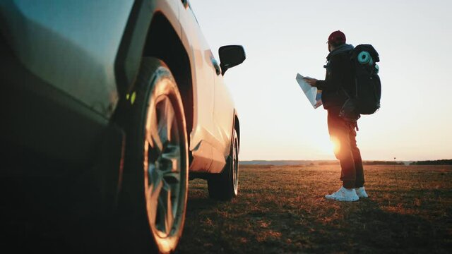 Silhouette tourist man reading map standing next to car during road trip. Hiker with backpack searches for a route on the map at sunset. Best adventure. Leisure, road trip, travel and people concept.