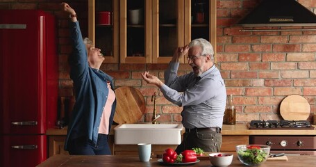 Happy loving sincere senior middle aged family couple dancing together in kitchen, enjoying energetic hobby weekend pastime, distracted from preparing healthy food, domestic activity concept.
