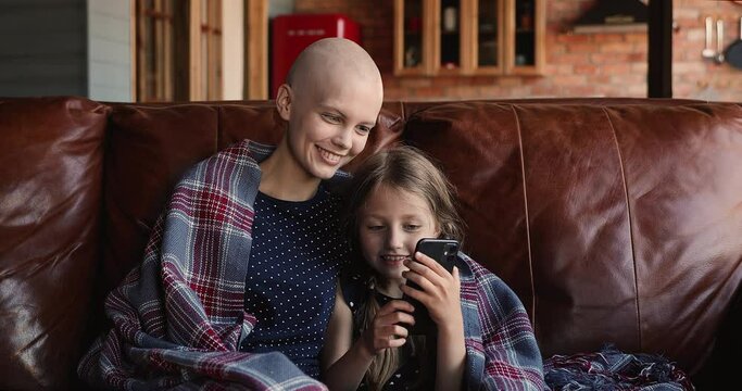 Happy young cured from cancer woman with bald head sitting with little kid daughter on sofa, covered in warm plaid, watching funny photo or video content in social network, using mobile applications.