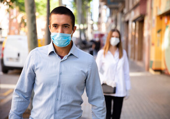 Focused adult man wearing medical mask walking along city street in sunny spring morning. New life reality during coronavirus pandemic.