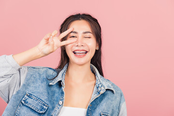 Asian happy portrait beautiful cute young woman wear denim smile standing showing finger making v-sign victory symbol near eye looking to camera studio shot isolated on pink background with copy space