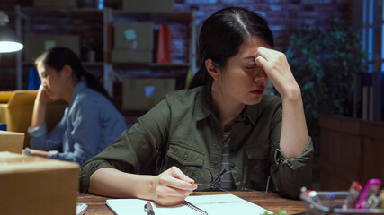 Start up small business freelance woman worker hard working overtime with colleague and feeling head painful. lady staff suffering headache in night office while coworker fell asleep leaning on desk.