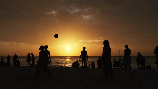 Silhouettes of people playing beach volleyball at sunset. Slow Motion. Zanzibar, Africa. Players on a sandy beach by the ocean among other vacationers. Orange sun with a halo setting on the horizon.