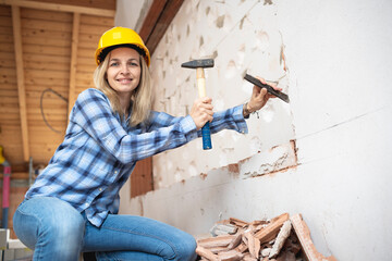 pretty young worker woman with blue work shirt and yellow protective helmet works on construction site and holds hammer in her hand, concept of female worker