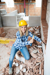 pretty young worker woman with blue work shirt and yellow protective helmet works on construction site and holds hammer in her hand, concept of female worker