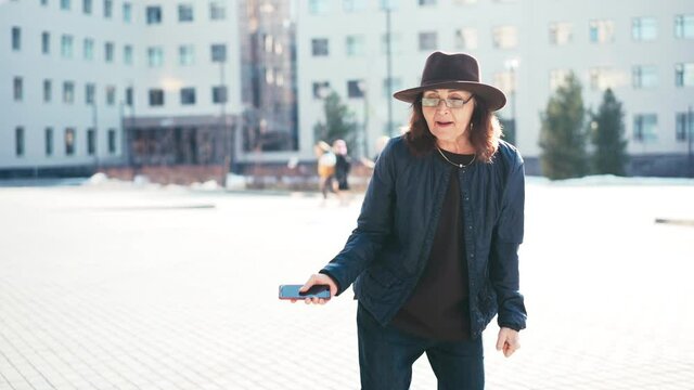 Real-time footage of an overjoyed senior woman in glasses dancing in the street on a sunny day with a smartphone in her hands.